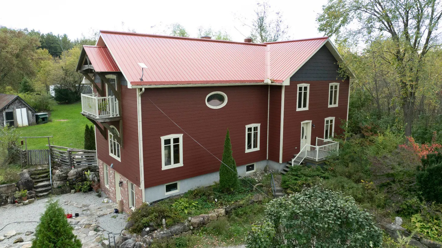 A two-story house with a red roof and maroon siding, surrounded by trees and greenery. A small balcony is visible on the left side.