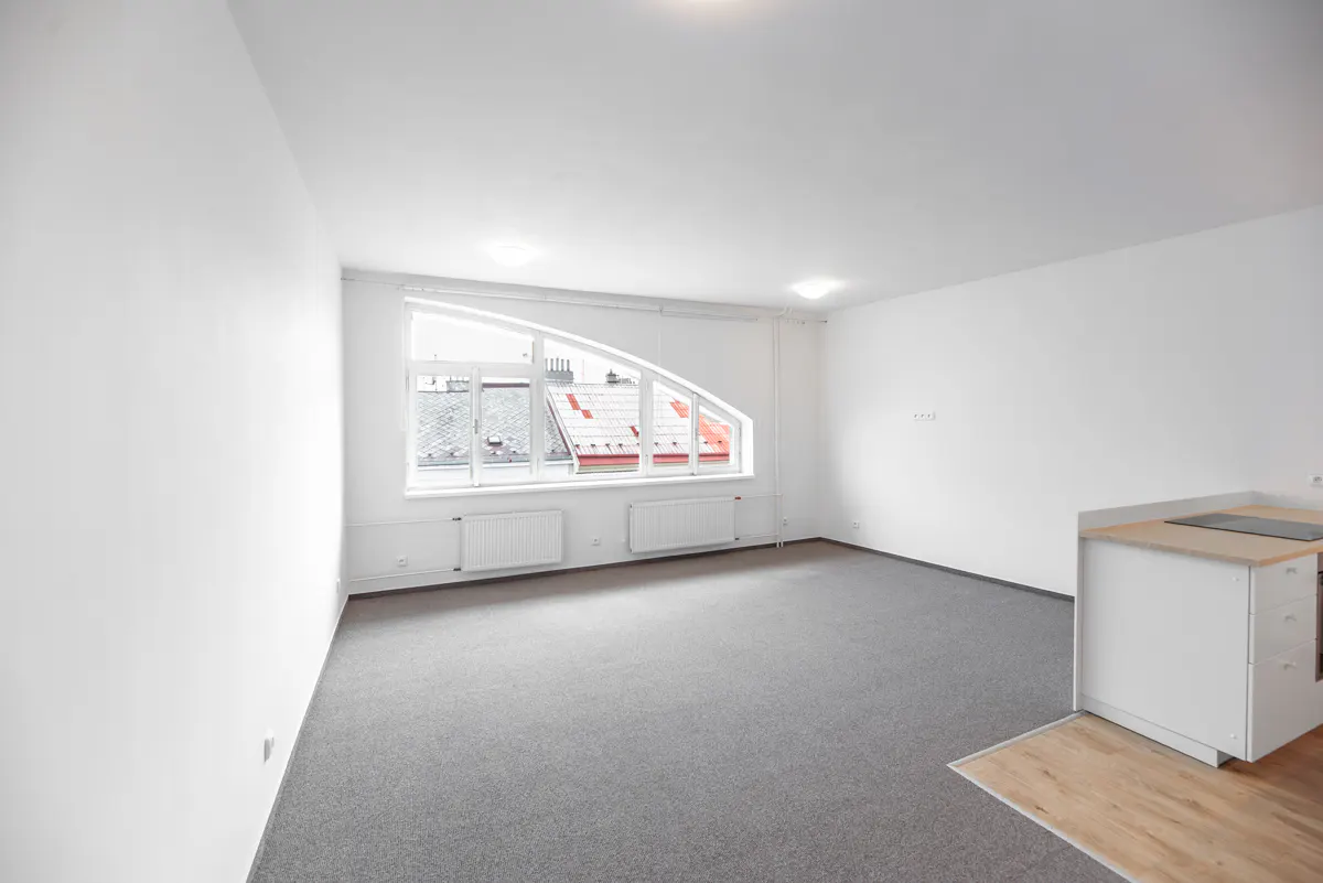 Bright, empty apartment with gray carpet, white walls, and a large, arched window overlooking rooftops. A small kitchen area is visible.