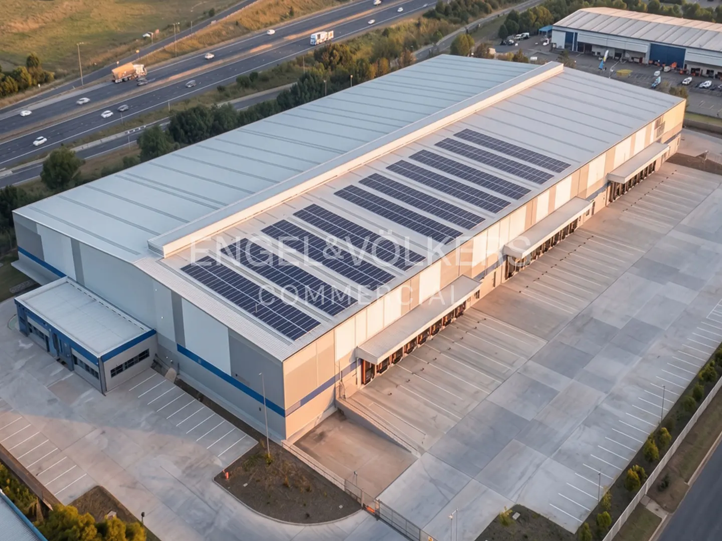 Aerial view of a large, modern warehouse with solar panels on the roof and a spacious parking lot.