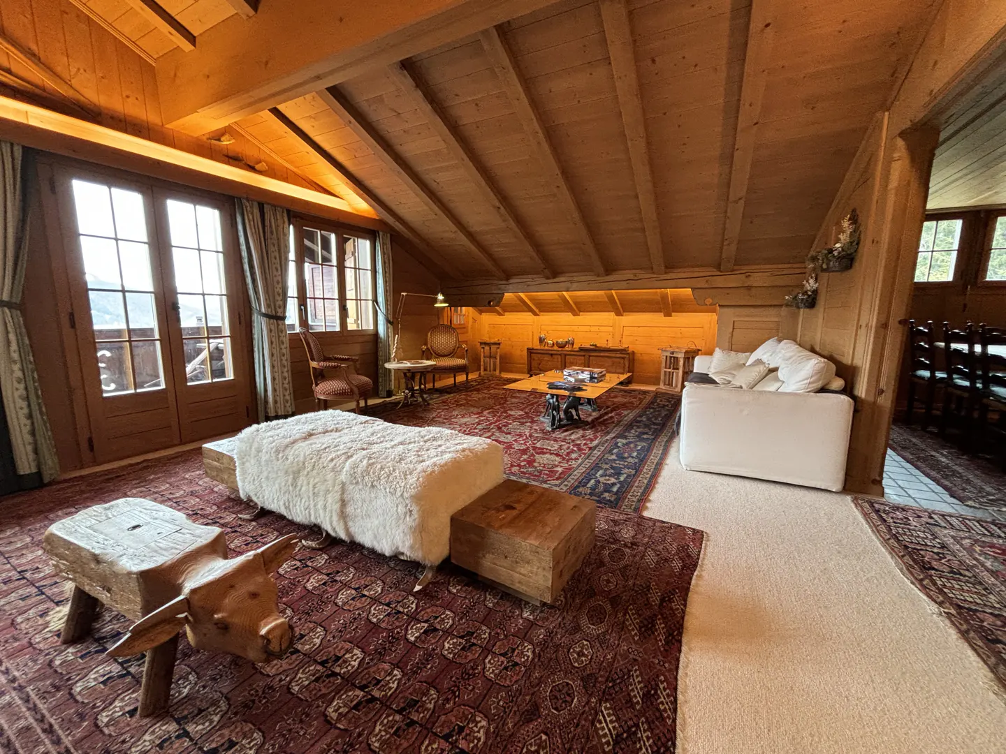 Attic living room with wood-paneled walls and ceiling, a red patterned rug, and a white sofa. A wooden cow stool sits in the foreground.