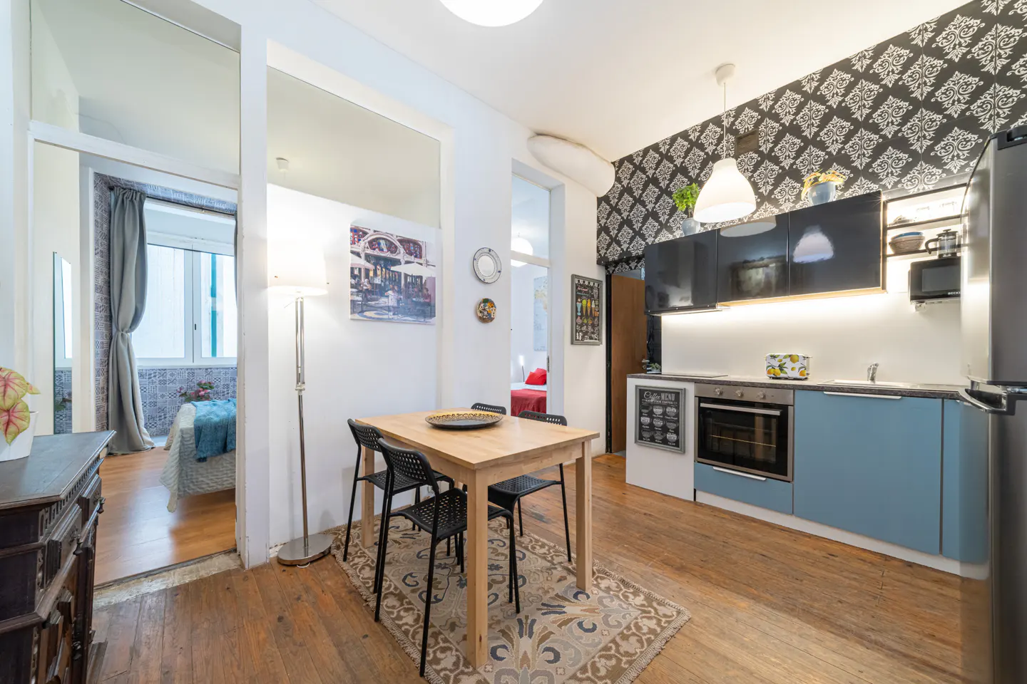 Apartment interior with a wood dining table, black chairs, and a blue kitchen with black cabinets. A bedroom is visible in the background.