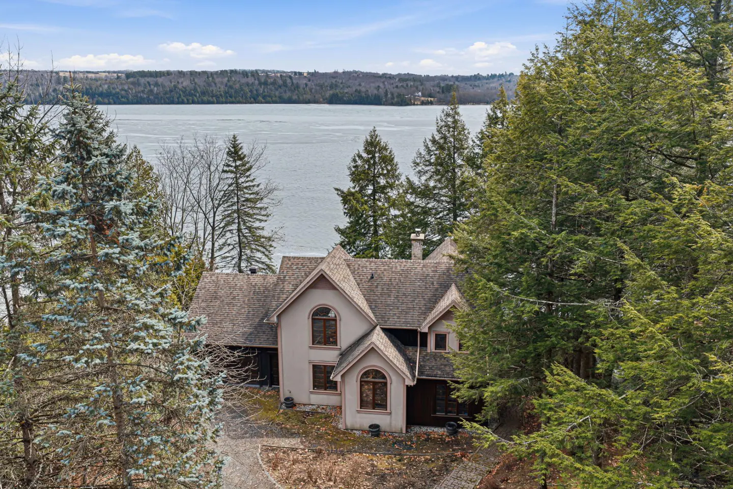 A beige house with a brown roof is surrounded by green trees, with a lake and blue sky in the background.