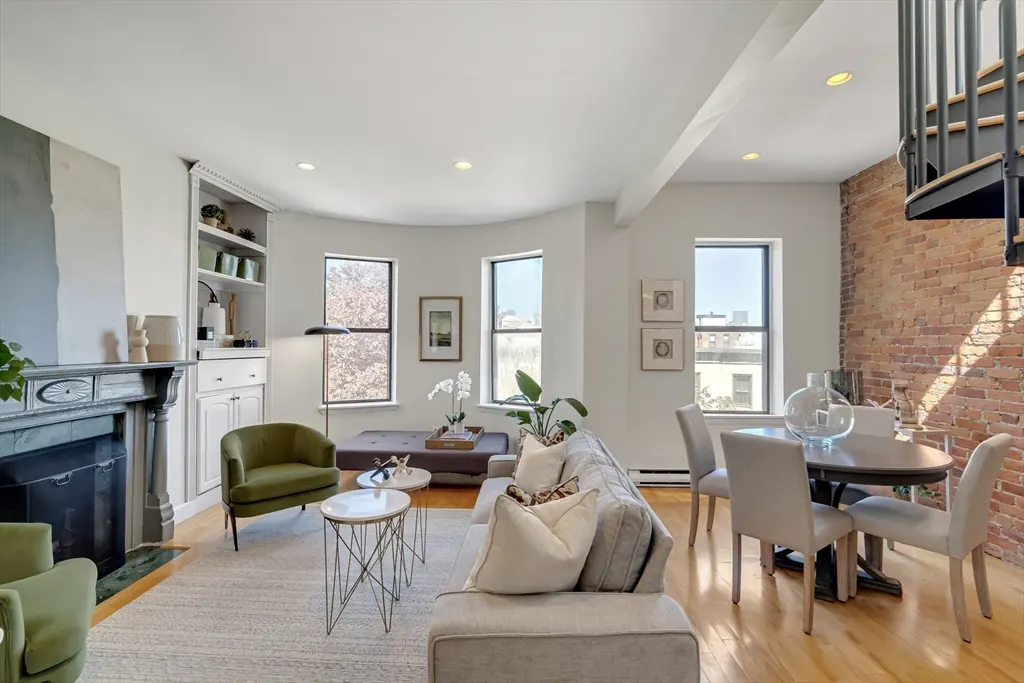 Bright living room with hardwood floors, a gray sofa, green chairs, and a dining table with a brick wall and spiral staircase.