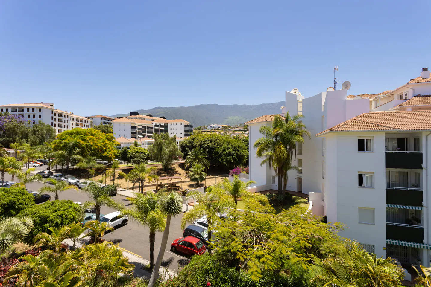 High-angle view of white apartment buildings with terracotta roofs, palm trees, a parking lot, and mountains in the distance.