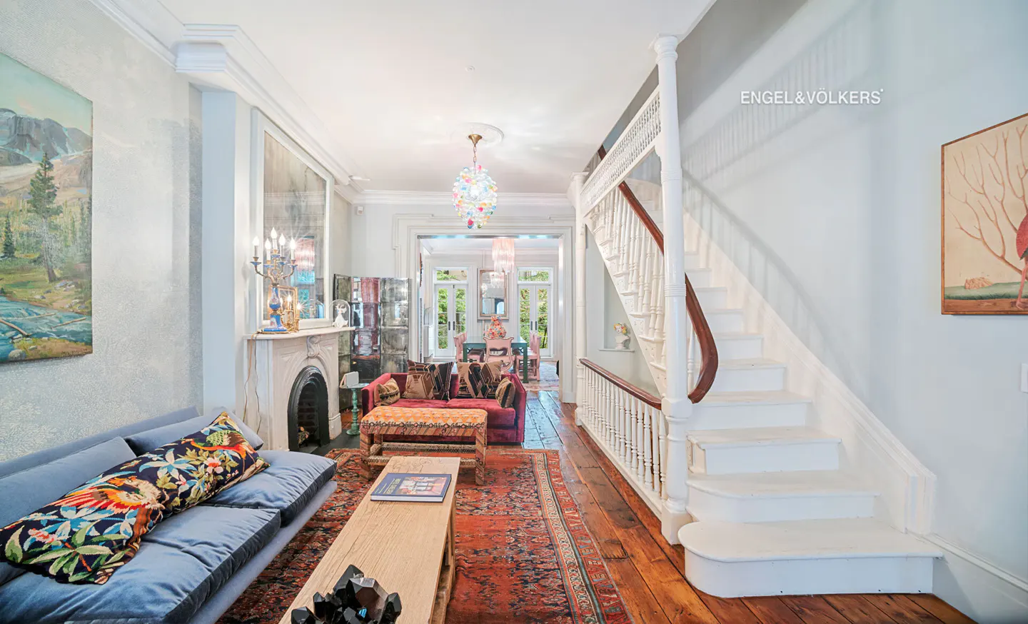 Elegant living room with a blue sofa, red patterned rug, white staircase, and a colorful chandelier over a dining area.