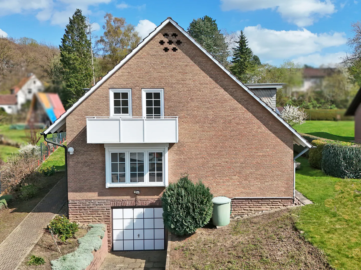 Brick house with white trim, balcony, and garage door. Green bushes and trees surround the property. Blue sky with clouds.