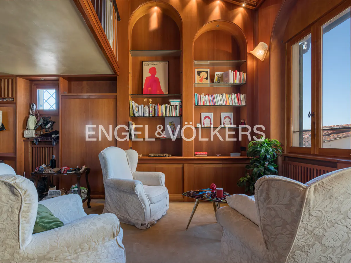 Living room with wood paneling, built-in bookshelves, and three white armchairs. A small table sits between two chairs. Window with blue sky.