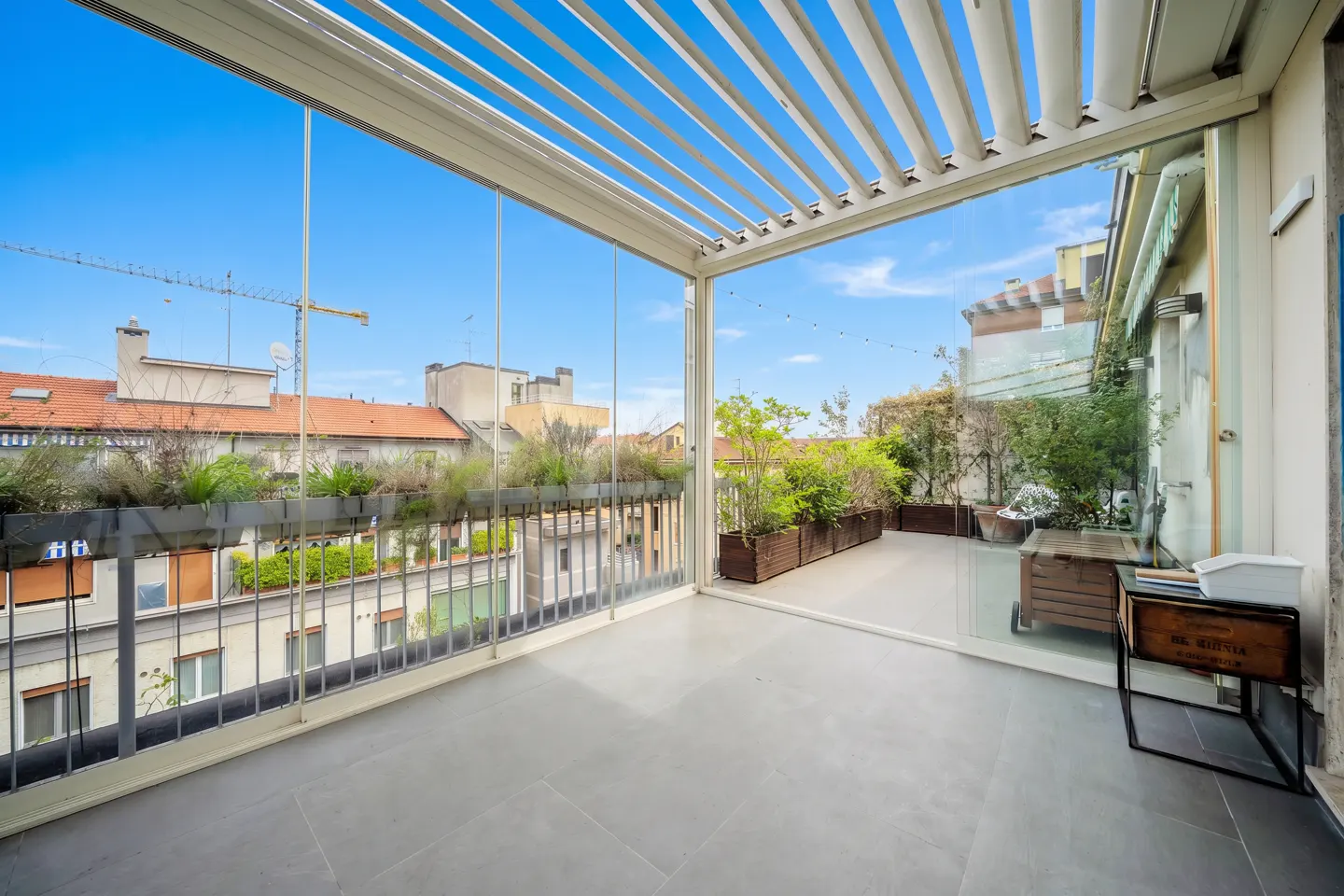 A modern balcony with a pergola roof, glass walls, and gray tile flooring. Outside, there are plants, buildings, and a blue sky.
