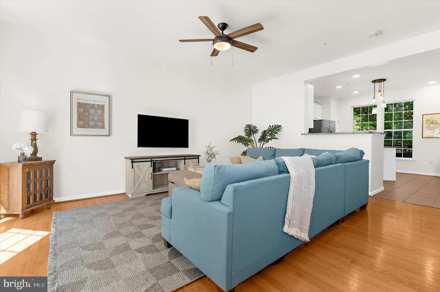 Bright living room with hardwood floors, a blue sectional sofa, and a ceiling fan. A TV sits above a sliding-door console.