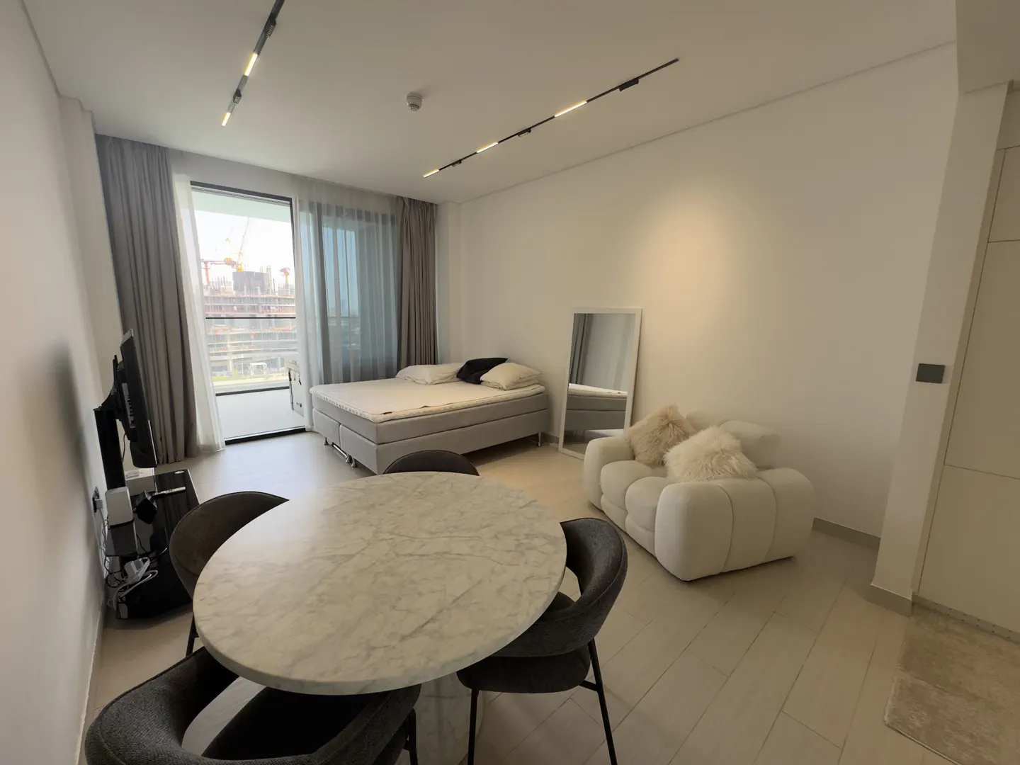 Bright studio apartment with a bed, sofa, and dining table. A large window shows a construction site.