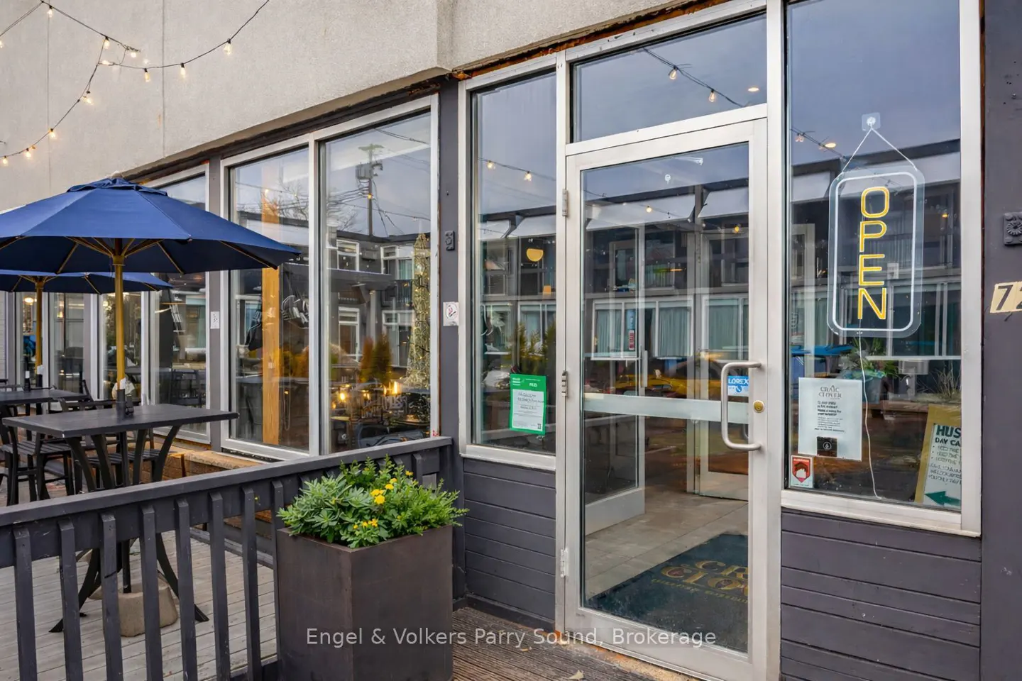 Exterior view of a restaurant with an "Open" sign in the window. Tables with blue umbrellas are on the patio. String lights hang above.