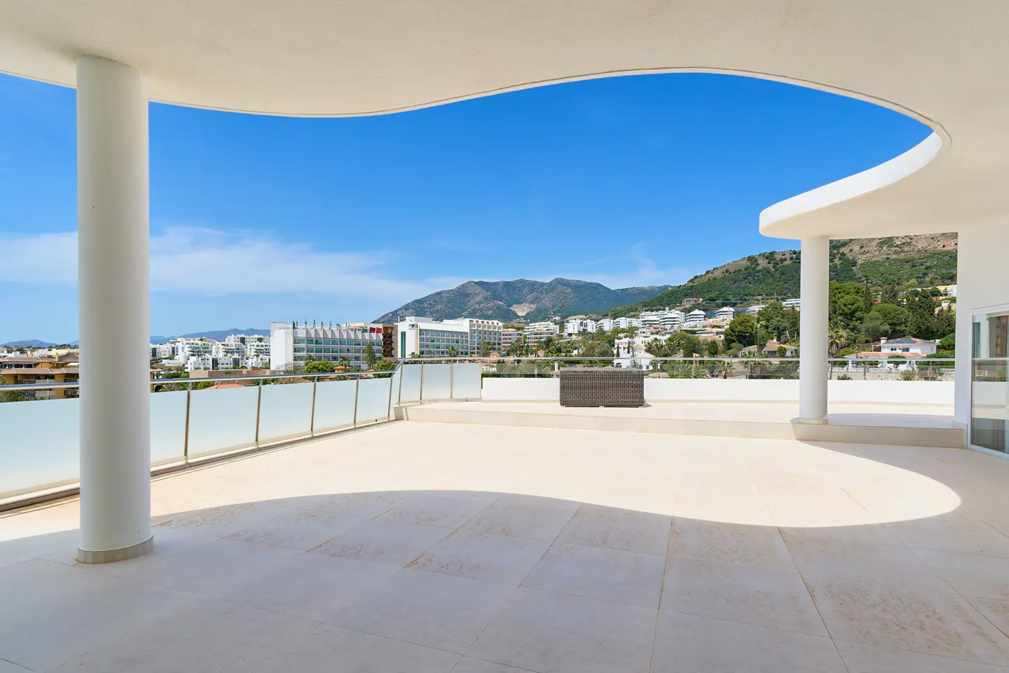Wide, sunlit balcony with white pillars and glass railings overlooking a city, mountains, and blue sky.