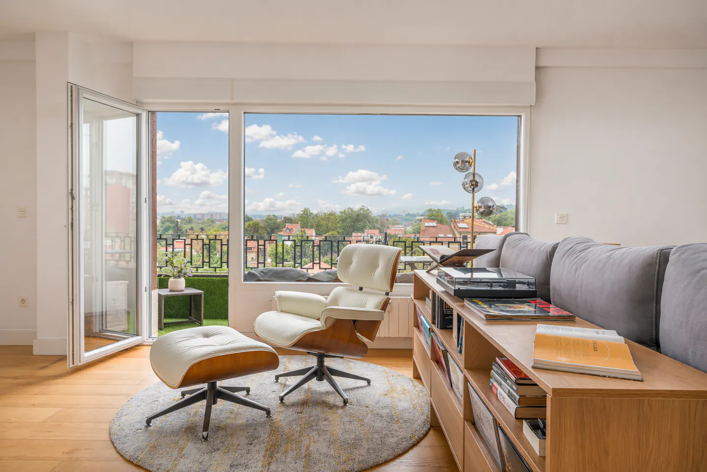 Bright living room with a white Eames chair and ottoman on a round rug, next to a bookcase and a large window with a city view.