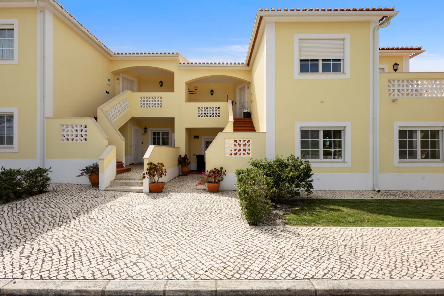 Exterior of a two-story yellow apartment building with white trim and a cobblestone driveway. Stairs lead to upper units. Potted plants decorate the entrance.