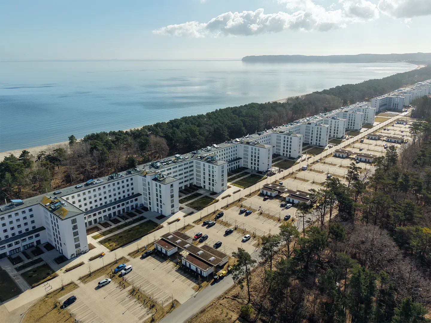 Aerial view of Prora, a long row of white buildings along the Baltic Sea coast, with parking lots and trees.