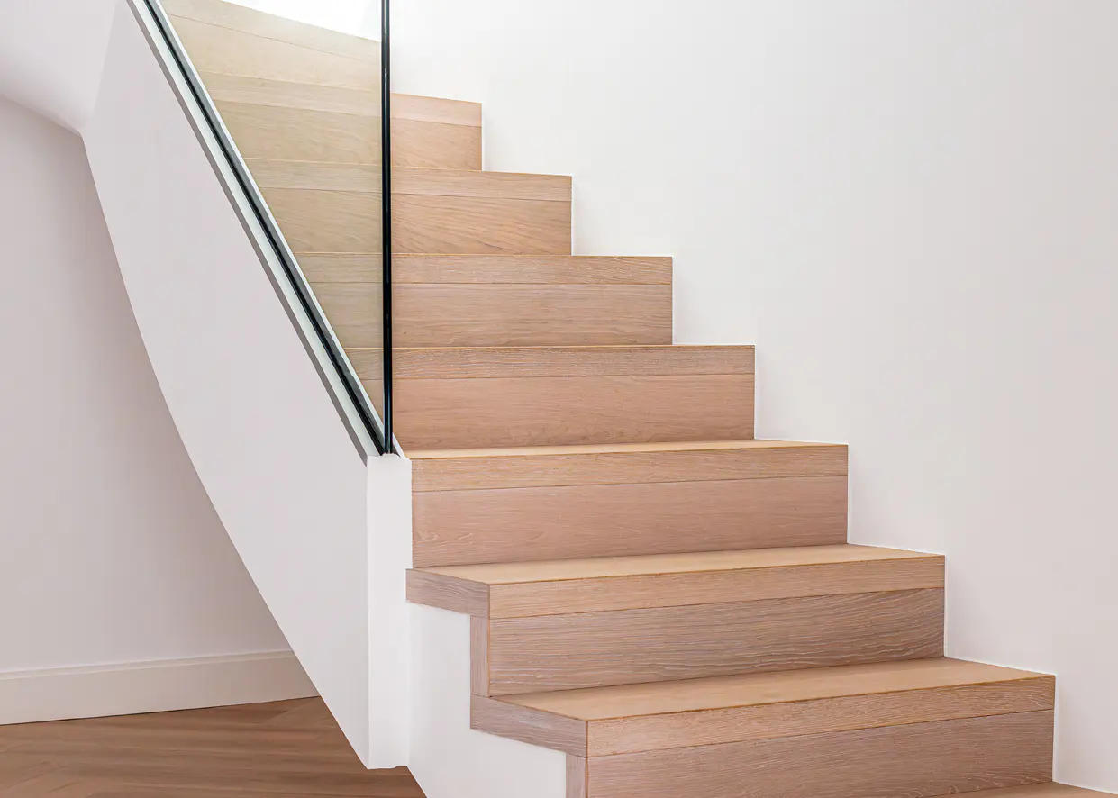 Modern staircase with light wood steps, white risers, and a glass railing against a white wall.