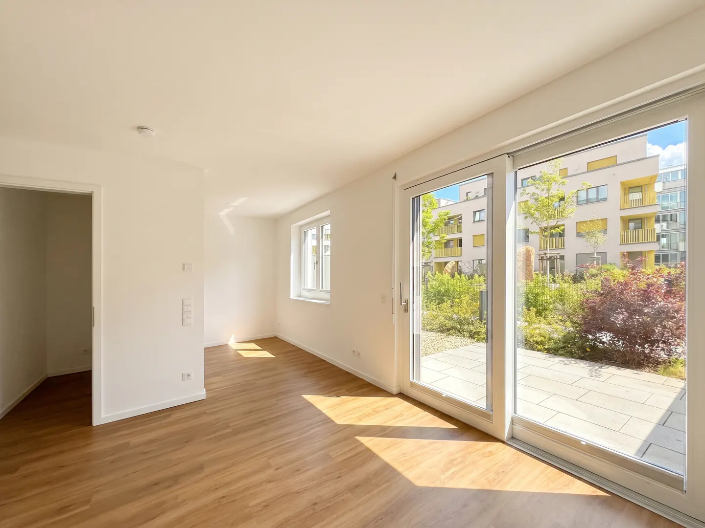 Bright, empty room with wood floors, white walls, and sliding glass doors to a patio and garden. A window and doorway are also visible.
