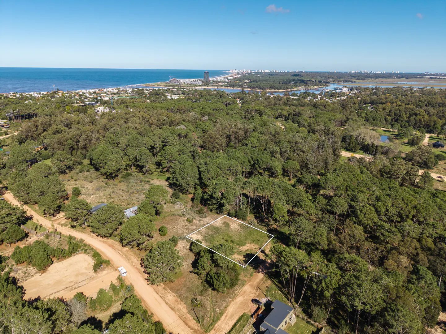 Aerial view of a vacant lot outlined in white, surrounded by green trees, with the ocean and city skyline in the background.