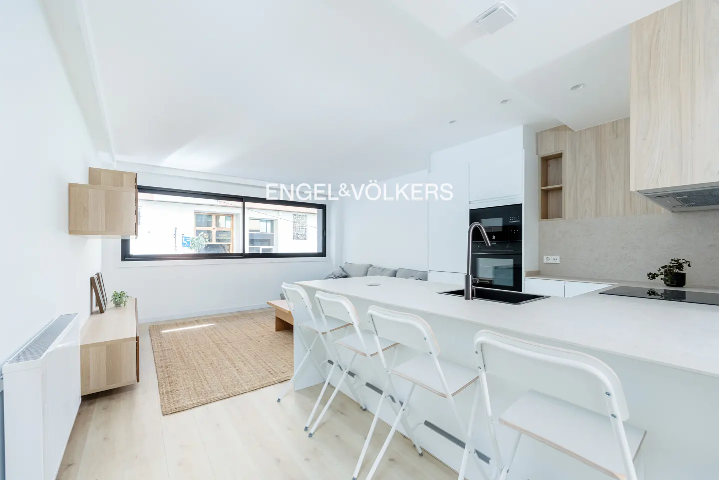 Modern open-plan kitchen and living area with white cabinets, a breakfast bar with white stools, and a light wood rug.