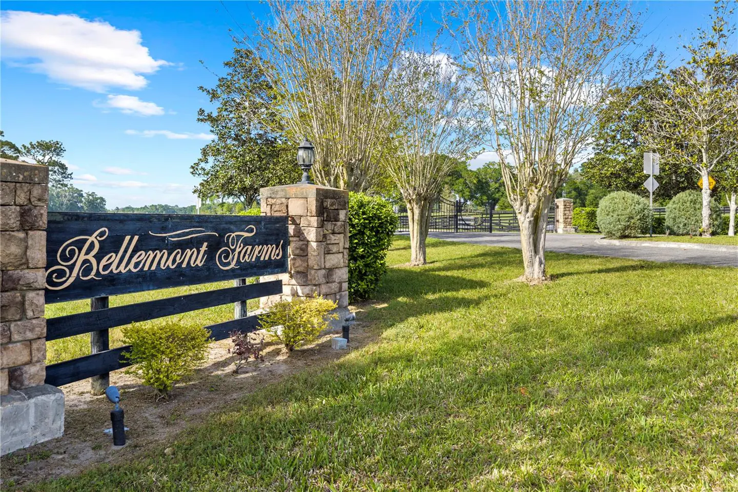 Entrance to Bellemont Farms with a black sign in gold lettering, stone pillars, and a black fence. Green lawn and trees under a blue sky.