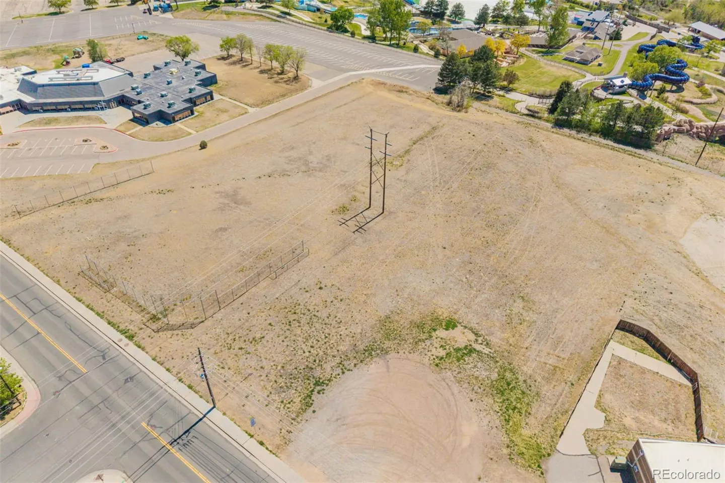 Aerial view of a large, vacant lot with dry, tan grass, utility poles, and a chain-link fence. A building and parking lot are visible in the background.