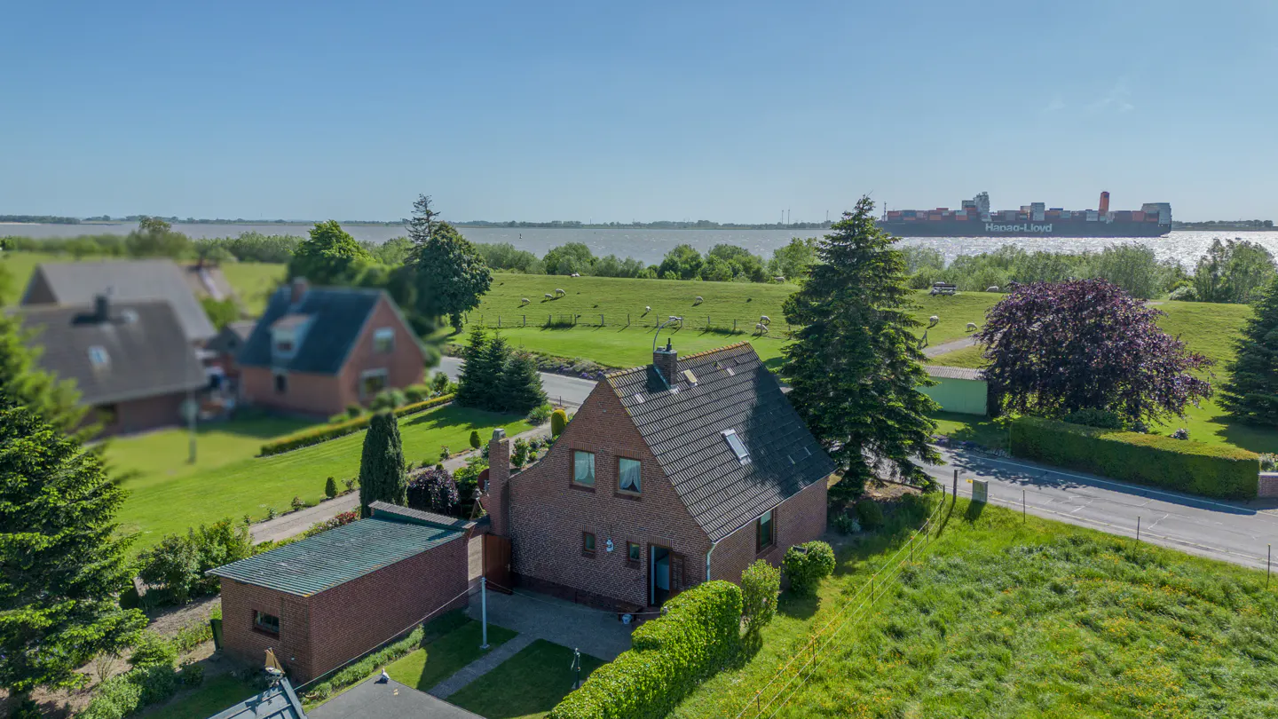 Aerial view of a brick house with a yard, near a grassy dike and a river with a large cargo ship in the background.