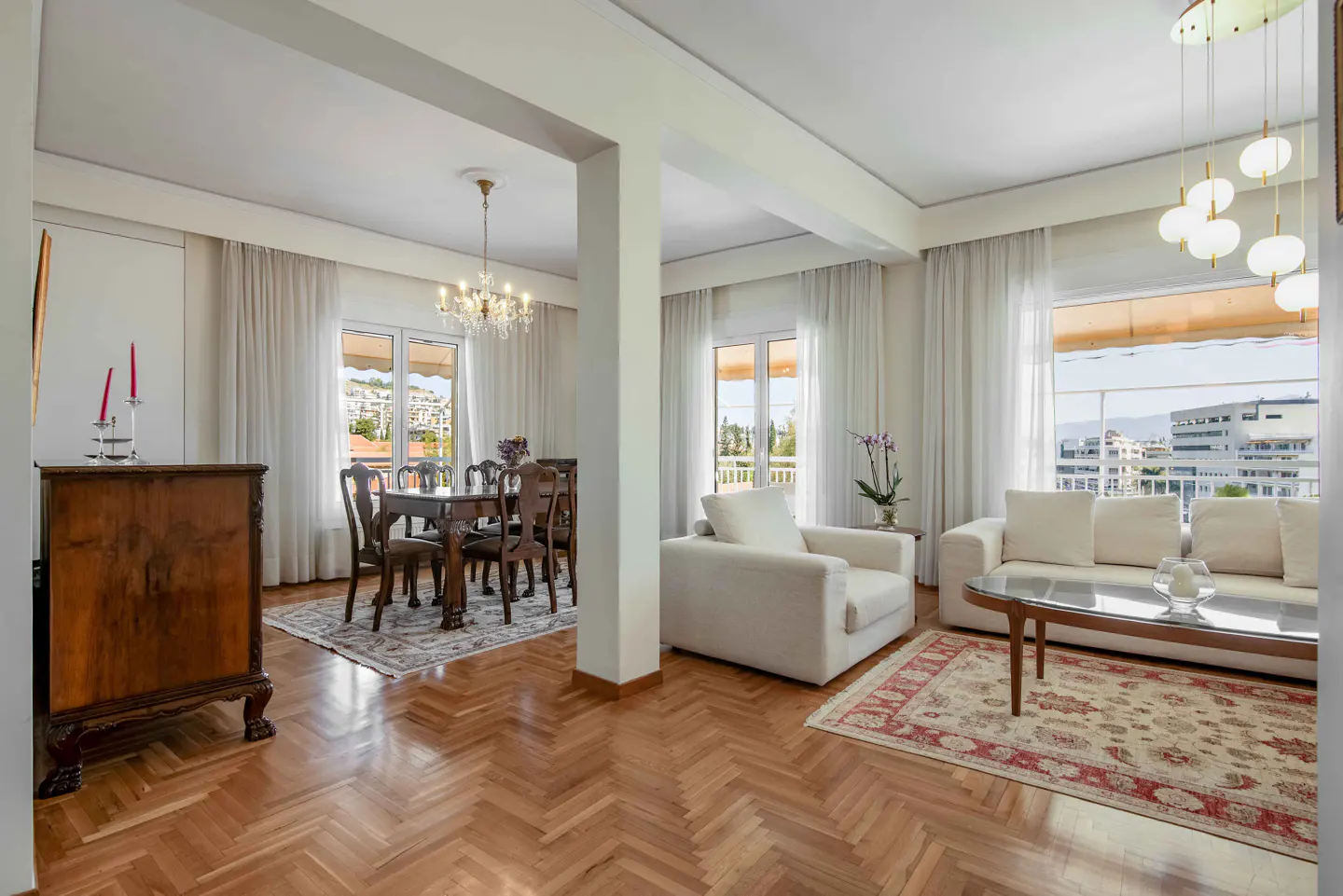 Bright, open-plan living and dining area with hardwood floors. White sofas, a glass coffee table, and a dining table with chairs are visible. Large windows offer city views.