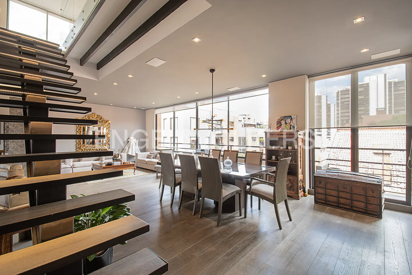 Open-concept living space with a dining table, floating stairs, and large windows. The room has hardwood floors and neutral-colored furniture.