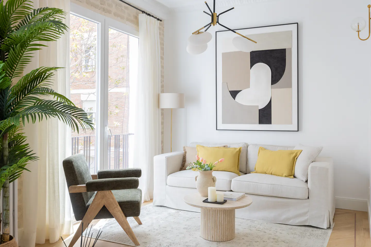 Bright living room with white walls, a sofa with yellow pillows, and a modern art print. A green chair and palm add a touch of nature.