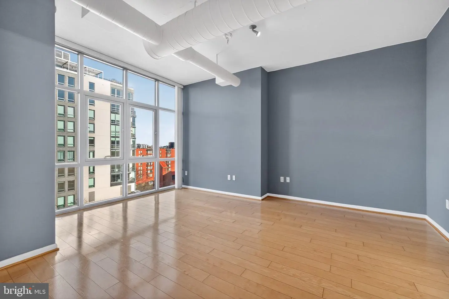 Empty room with hardwood floors, gray walls, and a large window showing city buildings. White ductwork is visible on the ceiling.