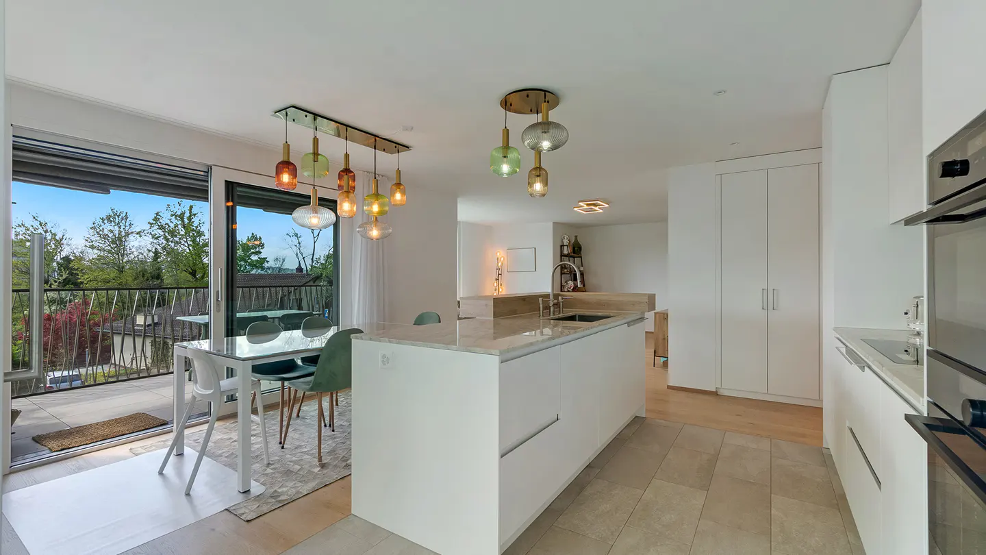 Bright, modern kitchen with white cabinets, island, and dining area. Colorful pendant lights hang above the table and island. Balcony with trees visible.