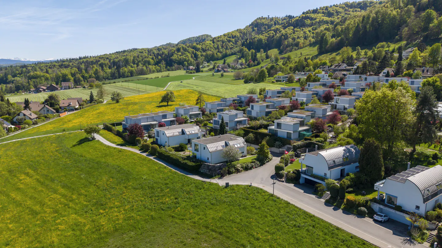 Aerial view of modern white houses nestled on a green hillside with trees and fields under a blue sky.