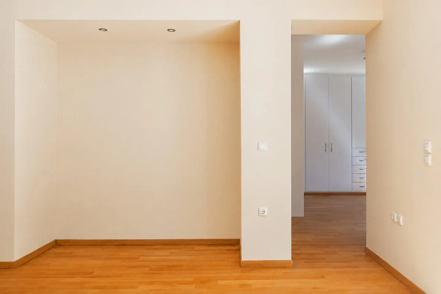 Empty room with light beige walls and wood floors. A doorway leads to a room with white closets.