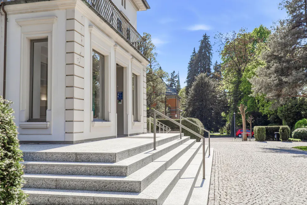 Exterior view of a white building with large windows and stone steps leading up to the entrance. A cobblestone path leads to the building.