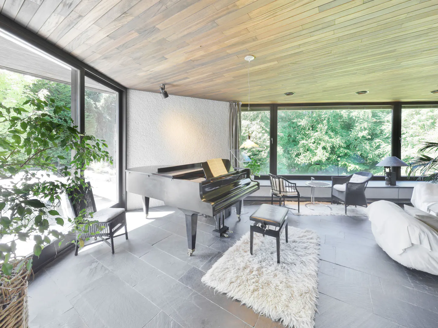 A black grand piano sits on a shaggy rug in a modern room with slate floors, wood-paneled ceiling, and floor-to-ceiling windows.