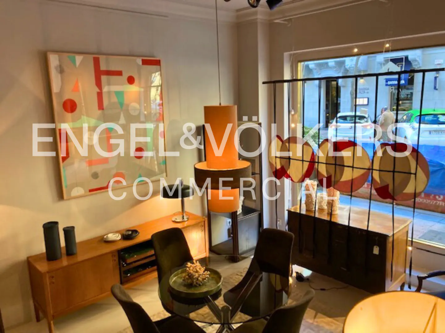 Interior view of a commercial space with modern decor, featuring a round glass table with chairs, a wooden cabinet, and an abstract art piece.