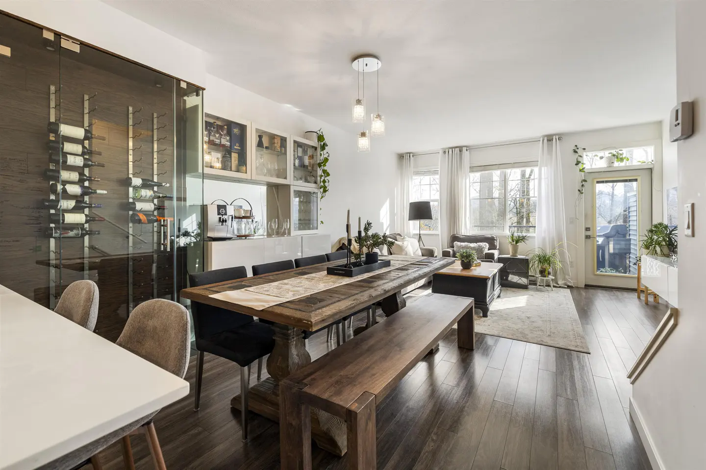 Bright, open-concept living space with dark wood floors. A large wooden table and bench sit near a glass-enclosed wine rack. Living room with sofa and windows in the background.