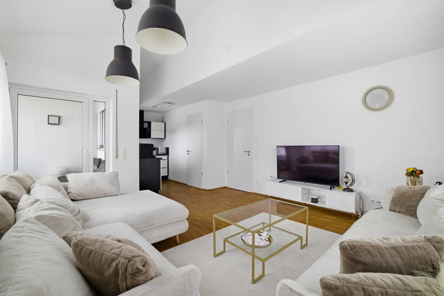 Bright living room with white walls, a large white sectional sofa, and a glass coffee table. A TV sits on a white console.