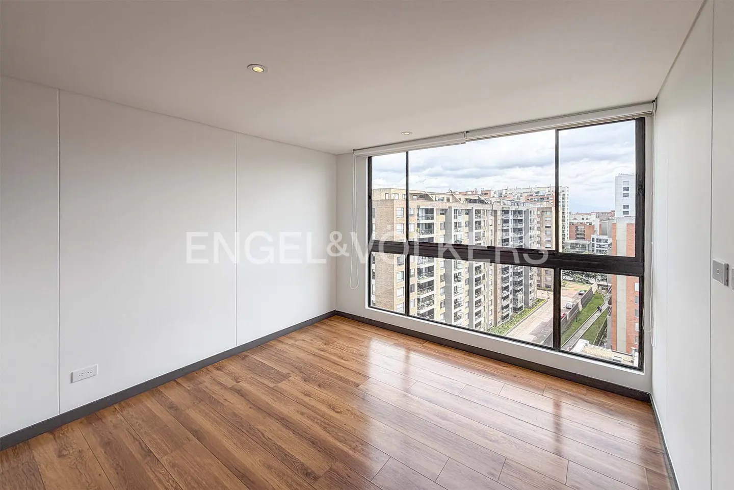 Bright, empty room with wood floors and white walls. A large window shows a city view.