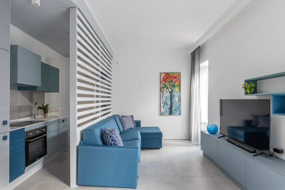 A modern apartment interior with a blue sofa, kitchen, and TV stand. A white slat wall divides the kitchen and living area. An abstract tree painting hangs on the wall.