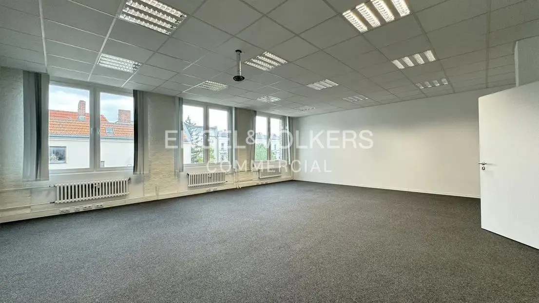Bright, empty office space with gray carpet, white walls, and large windows. Radiators are under the windows. Engel & Volkers logo on the wall.