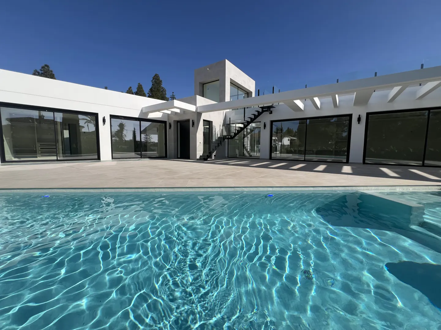 A modern white house with black framed windows overlooks a blue swimming pool on a sunny day.