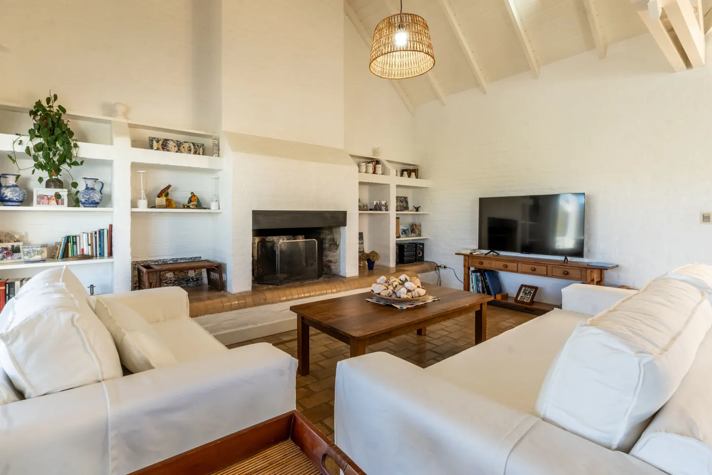 Bright living room with white walls, vaulted ceiling, and brick fireplace. Two white sofas face a wood coffee table and TV. Built-in shelves display decor.