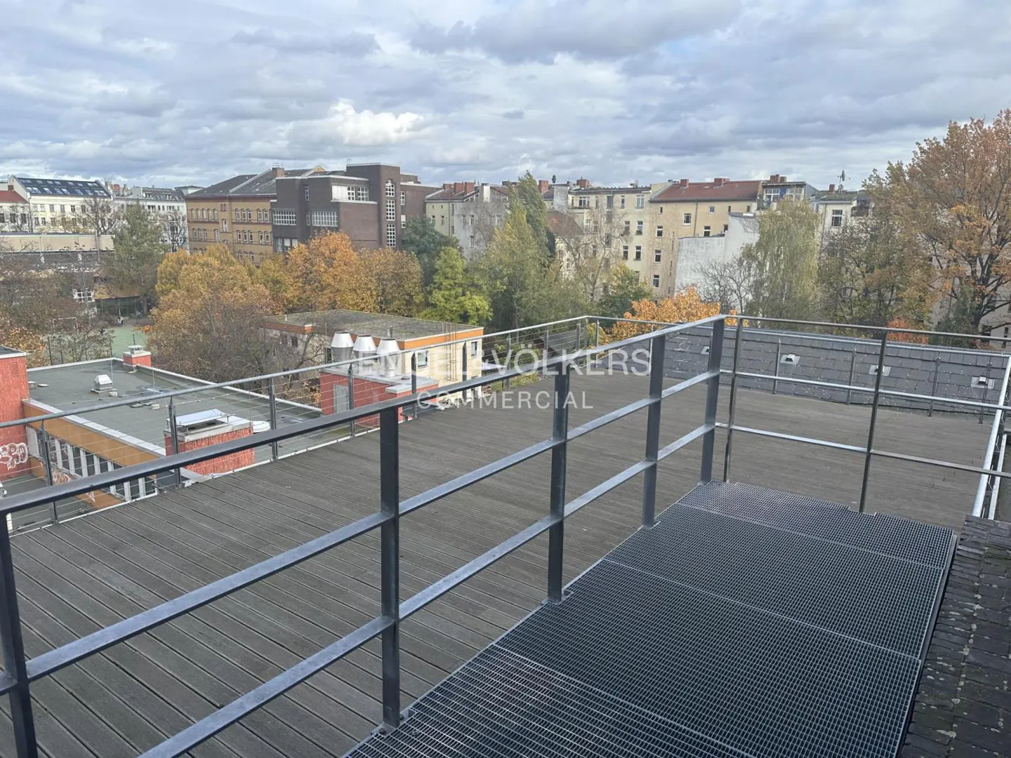View from a rooftop with metal railings, overlooking buildings and trees in autumn colors under a cloudy sky.
