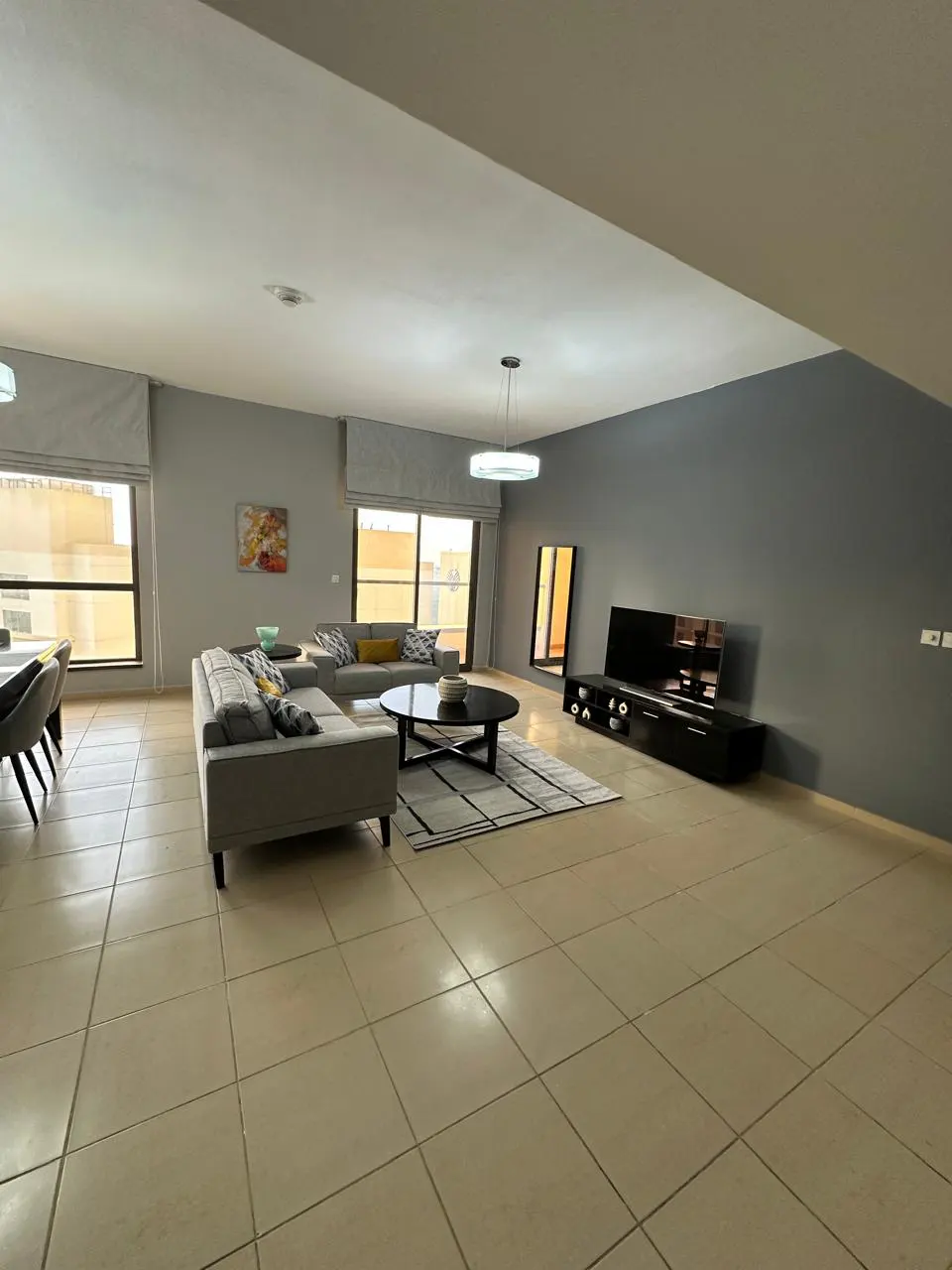 A spacious living room with gray sectional sofa, black TV stand, round coffee table, and beige tile flooring.