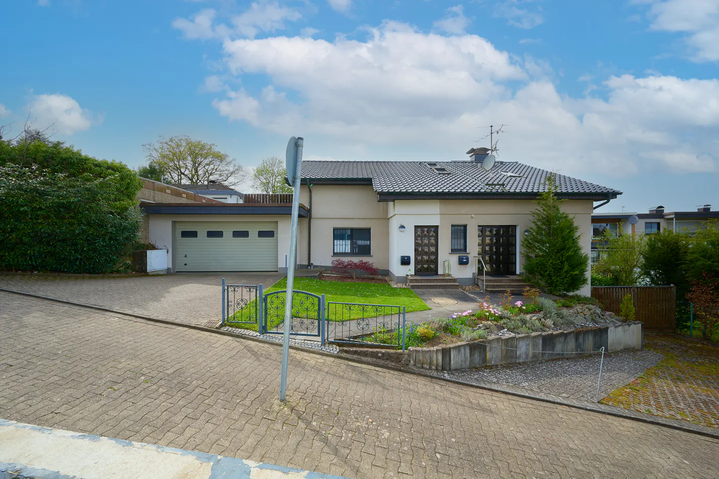 Beige two-story house with a gray roof, garage, and a small garden with flowers on a sunny day.