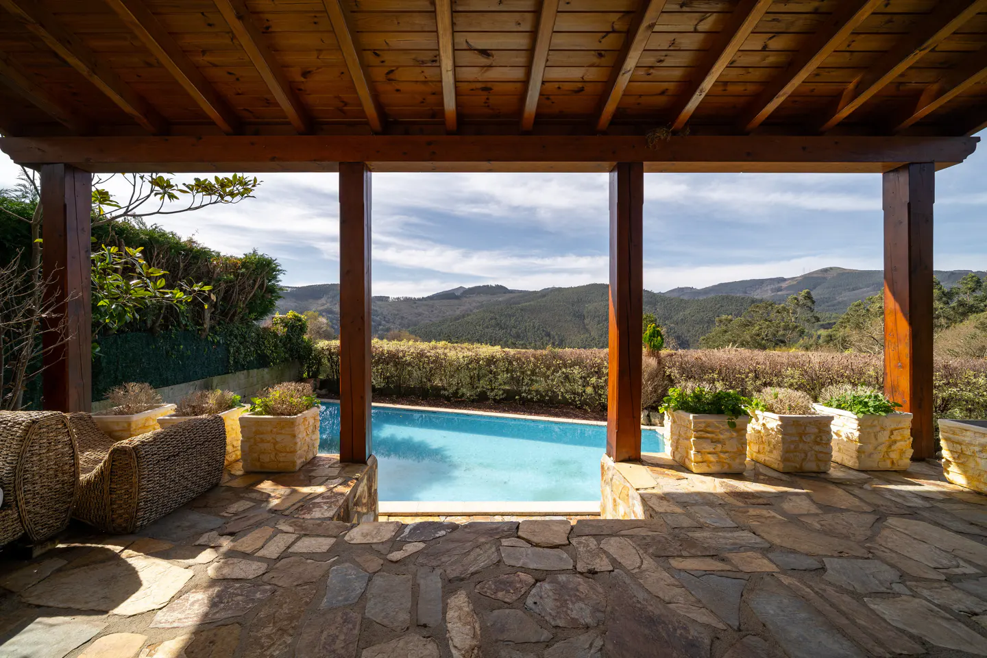 Covered patio with stone floor, wicker furniture, and potted plants overlooking a blue swimming pool and distant green hills.