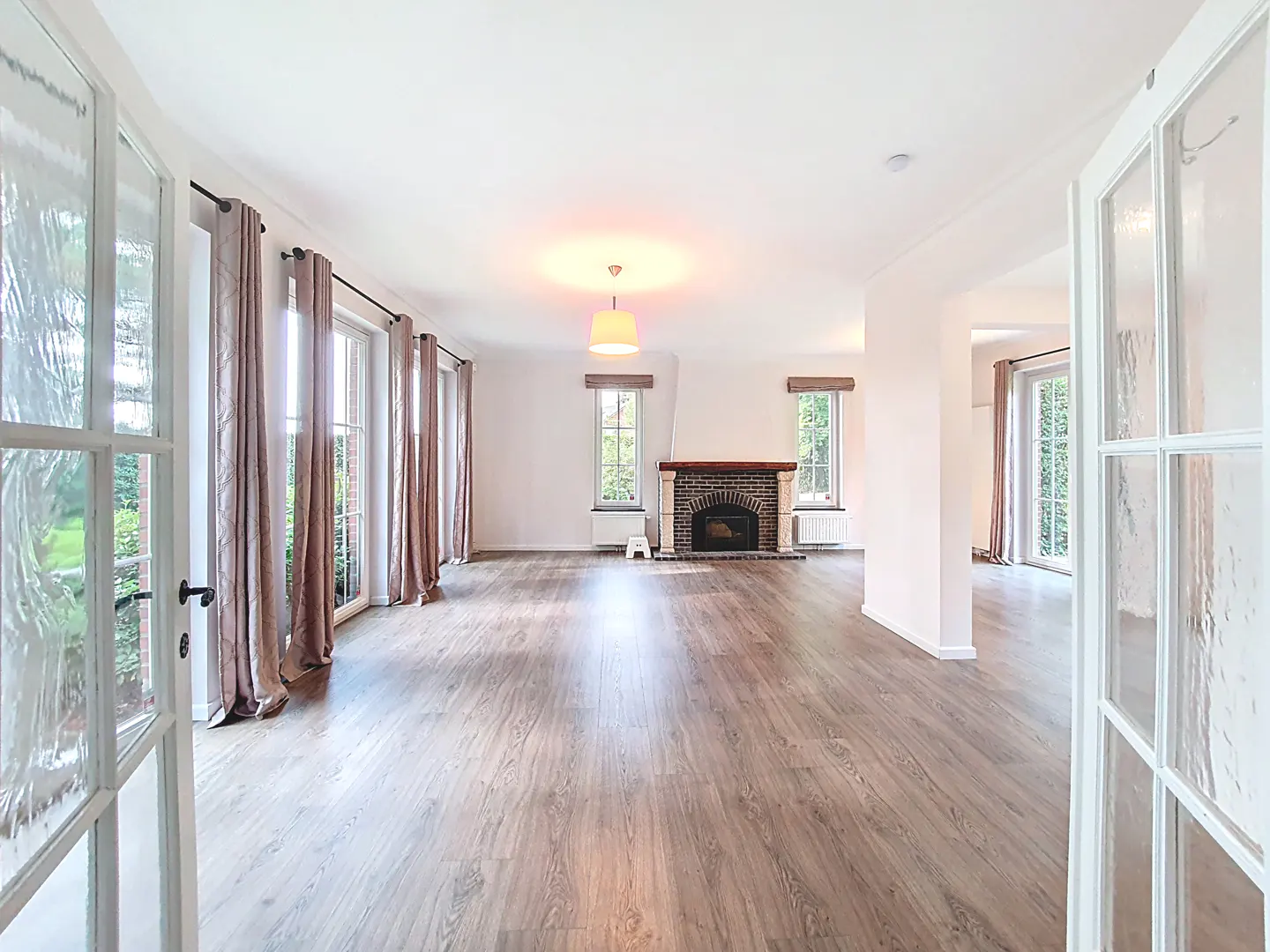 Bright, empty living room with wood floors, a brick fireplace, and white-framed windows with mauve curtains. Glass doors frame the view.