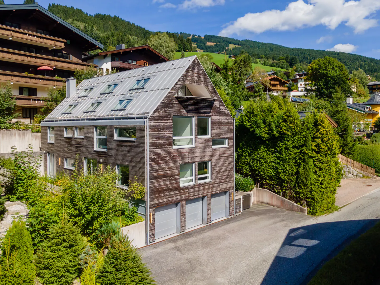 A modern, three-story brown house with a metal roof and multiple skylights, set against a backdrop of green trees and mountains.