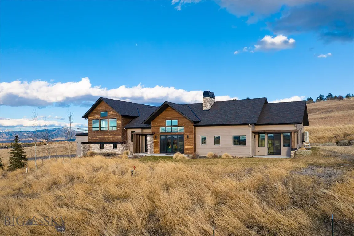 A modern home with wood and stone accents sits in a field of tall, dry grass under a blue sky with scattered clouds.